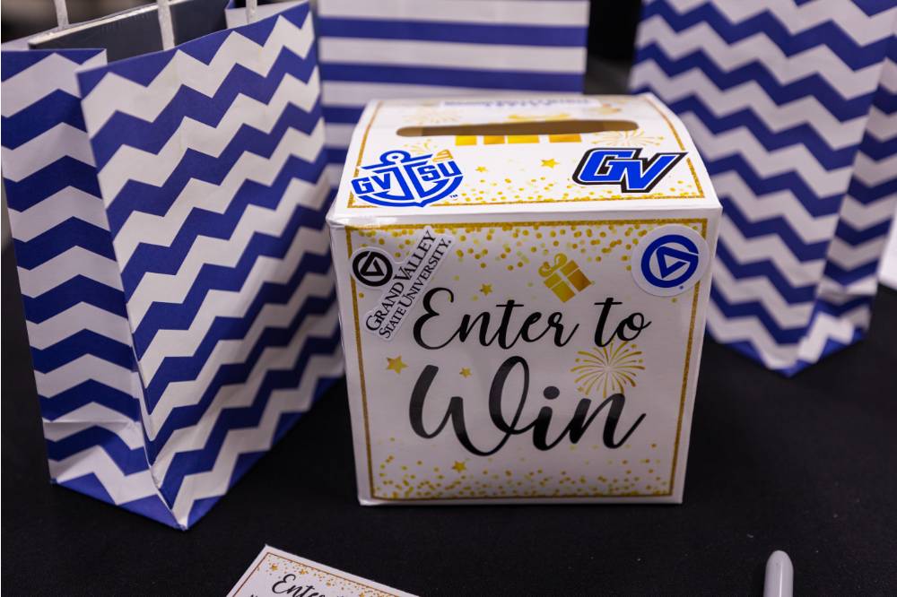"Enter to Win" Raffle box decorated with GVSU stickers and three blue and white striped bags